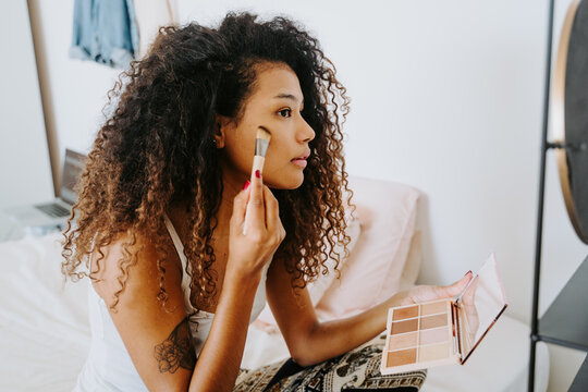 Side view of young curly haired ethnic female applying blush from palette on cheek with cosmetic brush during daily makeup procedure at home