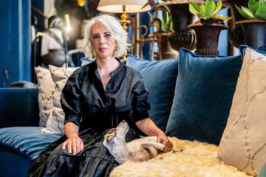 Charming Mature Female In Elegant Black Dress Sitting On Comfortable Couch And Stroking Purebred Dog While Relaxing In Stylish Living Room With Vintage Decor