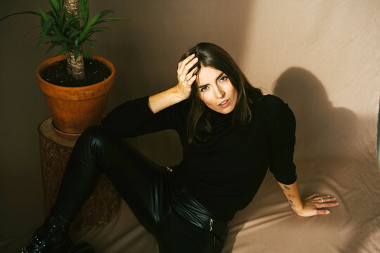 Confident young tattooed female in stylish black leather pants and turtleneck sitting on floor near potted plant and looking at camera in light studio