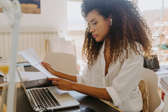 Side view of female bookkeeper with true wireless earbuds sitting at table in home office and analyzing financial reports and paper documents during remote work