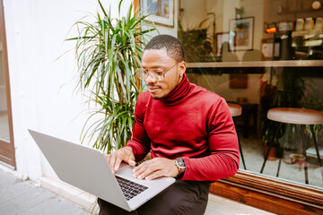 Rich stylish focused African American male entrepreneur sitting on bench near cafe outdoors and browsing laptop while working remotely on project