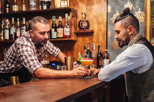 Side view of middle aged barkeeper giving cocktail with raspberries to elegant bearded man while working in bar