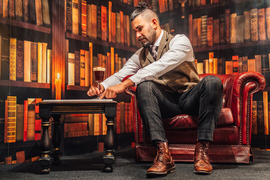 Full body of man in elegant suit holding glass of Irish coffee while resting in comfortable armchair