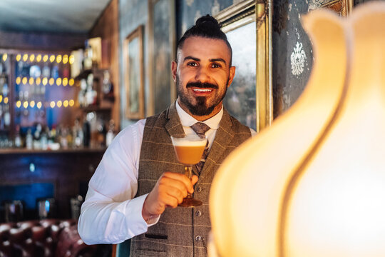 Cheerful bearded man holding glass of Irish coffee standing near old fashioned lamp in classic room looking at camera