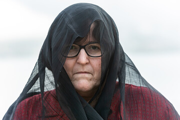 portrait of a middle-aged lady with a black headscarf on her head. Duel. In the background is the...