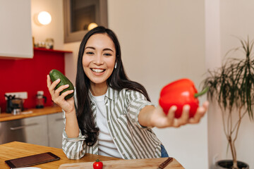 Positive woman shows red pepper and holds avocado while posing in kitchen