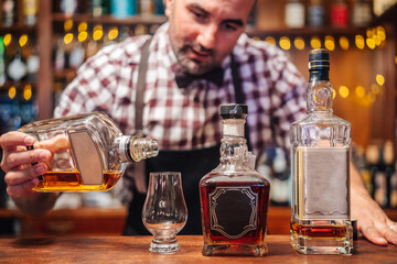 Crop brutal male bartender in apron pouring brandy in glass placed on counter with bottles of alcoholic drinks