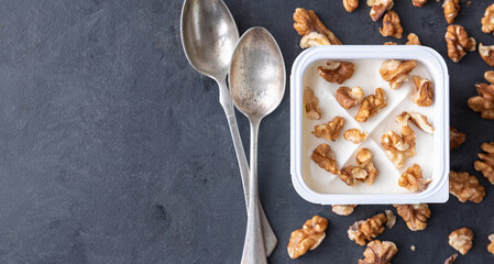 White yogurt in plastic square cup with marmalade and spoon in with walnuts on gray background. Flat view.