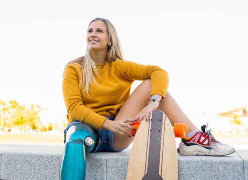 Carefree female skater in summer clothes and with bionic leg prosthesis sitting on stone border in city and looking away