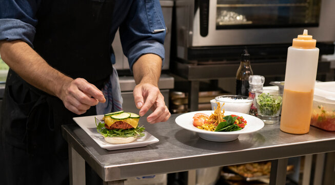 Crop unrecognizable male cook adding sliced fresh cucumber to hamburger with meat cutlet and cheese while preparing snack in restaurant kitchen
