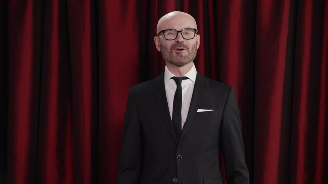 Late-night talk show host is performing his monologue against red curtain, looking into camera. TV broadcast style show. Shot with RED cinema camera