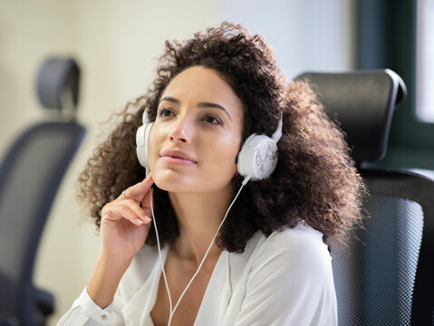 Young thoughtful Hispanic female employee in white blouse and headphones looking away while working at table with computer in modern workplace