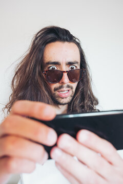 Overwhelmed young long haired hipster guy in sunglasses looking at screen of smartphone with astonished face expression while getting incredible news