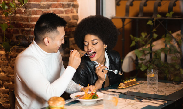 Happy Asian man feeding cheerful African American girlfriend with salad while having fun during lunch in cafe