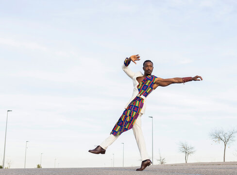 Side view of African American male dancer in trendy outfit performing energetic movements against blue sky