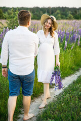 young beautiful couple a girl is pregnant in a white dress and a hat a man in a white shirt are walking along the field with lupins at sunset