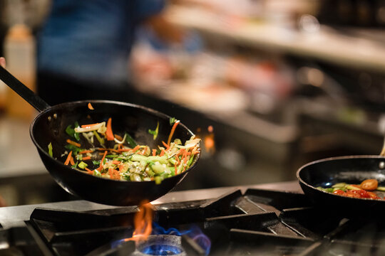 Skillet pan with fried vegetables being stirred by anonymous cook during preparation on gas stove in restaurant kitchen
