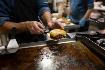 Crop anonymous cook with spatula taking roasted meat cutlet with melted cheese while preparing hamburger in restaurant kitchen