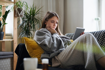 Ill young woman wrapped in blanket on sofa at home, using tablet.