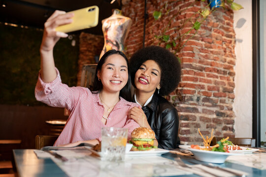 Playful young diverse female friends sitting at table with various dishes and taking selfie on smartphone with funny faces while having dinner together in restaurant