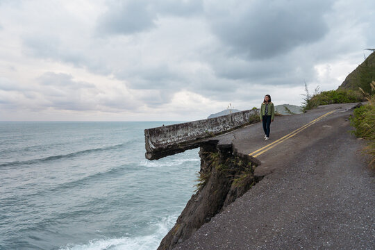 Unrecognizable distant asian ethnic female traveler standing on edge of destroyed asphalt road running along rough rocky coast near waving sea in overcast weather