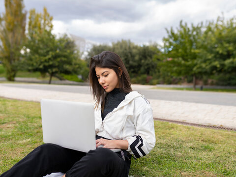 Modern young ethnic female student in casual wear sitting on green lawn and browsing laptop while spending time in park