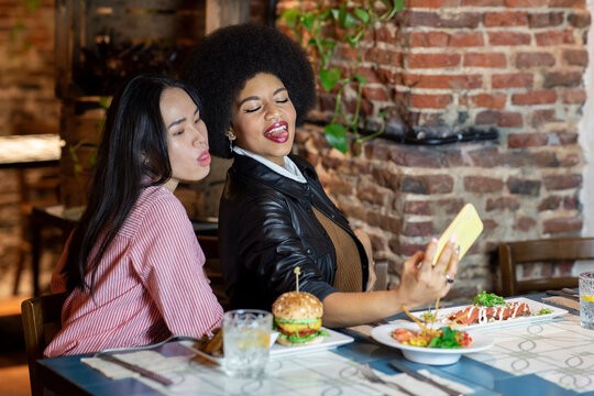 Playful young diverse female friends sitting at table with various dishes and taking selfie on smartphone with funny faces while having dinner together in restaurant