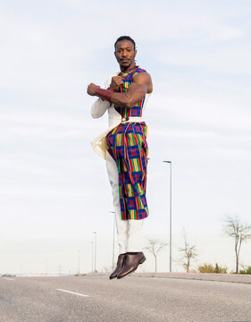 African male dancer in trendy outfit levitating above road while performing energetic movements against blue sky making arms gesture looking at camera