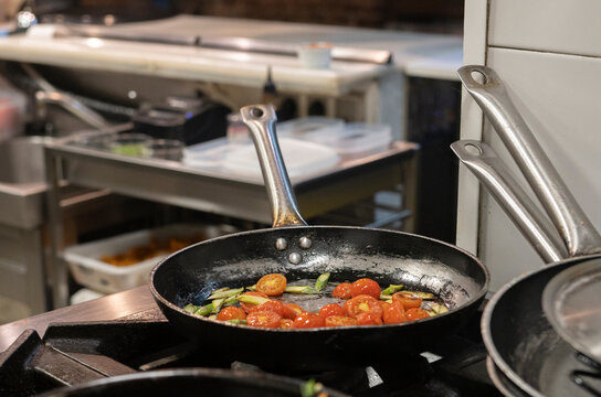 Skillet pan with fried tomatoes and greens placed on stove in modern restaurant kitchen during meal preparation