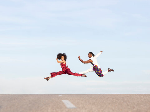 Side view of African American couple in trendy traditional clothes jumping high above asphalt road and performing dance on background of blue sky