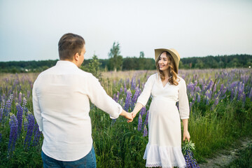 young beautiful couple a girl is pregnant in a white dress and a hat a man in a white shirt are walking along the field with lupins at sunset