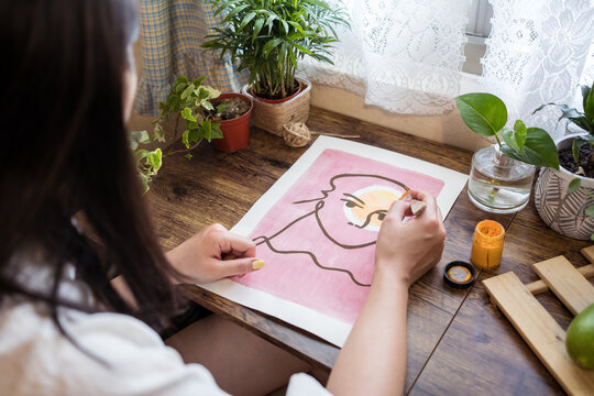 High angle side view of unrecognizable Asian female artist sitting at wooden table and creating picture on paper with paint and brush
