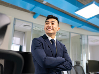 Cheerful young Asian male manager in formal suit looking away while standing near table with computers in contemporary coworking office