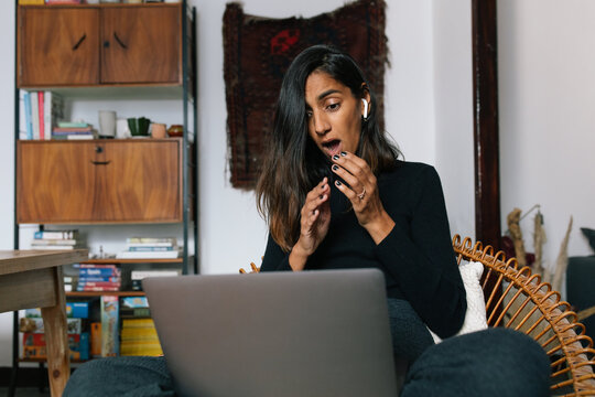 Low angle of astonished Indian female sitting with laptop and reading news online while covering mouth