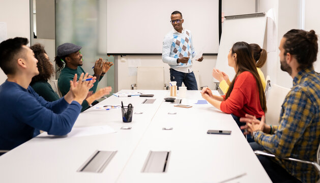 Cheerful multiracial coworkers sitting around table and applauding to male speaker during presentation and discussion of business strategy in modern workplace