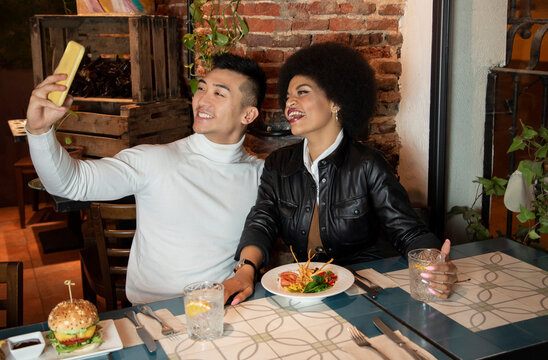 Happy young Asian man and African American girlfriend having fun and taking selfie on smartphone during lunch in restaurant