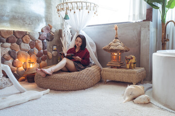 Full body of calm young Asian female traveler in casual clothes sitting on wicker pouf in stylish room with stone walls and typical oriental decorations while chilling in hotel in Taiwan