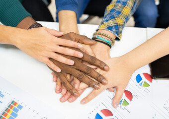 From above of crop anonymous multiracial colleagues in casual clothes gathering around table with charts and stacking hands together while working on business development plan in workplace