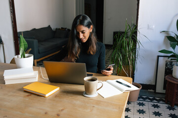 Focused Indian female freelancer sitting at table and typing on laptop while working on project in cozy home office