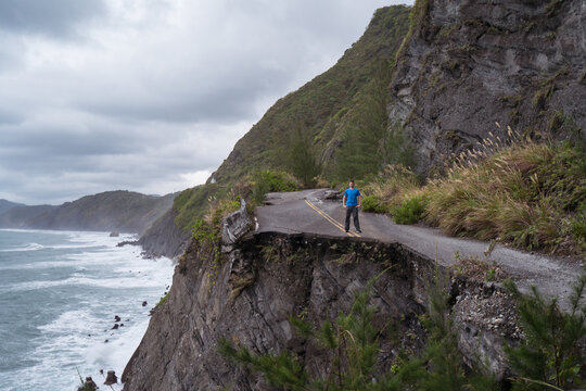 Unrecognizable distant male traveler standing on edge of destroyed asphalt road running along rough rocky coast near waving sea in overcast weather
