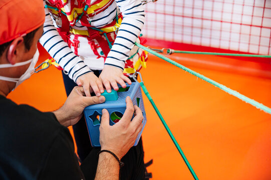 Crop gittle girl with Angelman syndrome playing with development game while being fixed with elastic bands during therapy session with anonymous caregiver