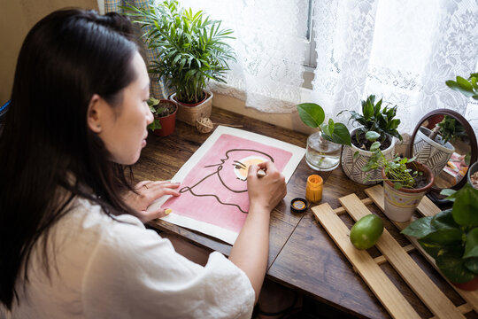 High angle side view of Asian female artist sitting at wooden table and creating picture on paper with paint and brush