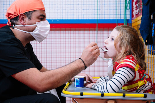 Side view of male specialist in medical mask giving yogurt to disable little girl wit Angelman syndrome while having break during developing activities