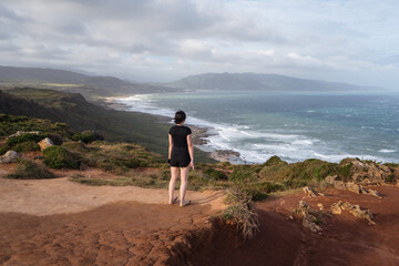 Full body back view of unrecognizable female traveler in black clothes standing alone on hilly shore against waving sea in overcast weather and contemplating nature