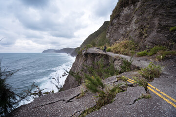 Distant unrecognizable couple of travelers in activewear strolling on half destroyed asphalt road on coastline in overcast weather with storming sea