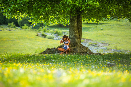 Full body of happy sensual young female in stylish swimwear touching long hair and smiling while relaxing on blooming grassy meadow near tree in Fuente De