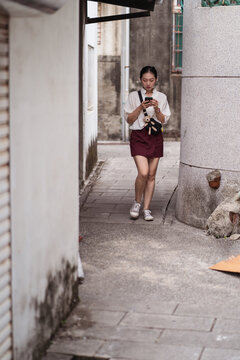 Full body of young Asian female traveler in casual clothes browsing mobile phone and checking map while exploring narrow street with stone buildings in Tainan city in Taiwan