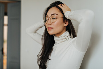 Brunette girl in glasses and sweater poses against white wall in apartment and massages her head