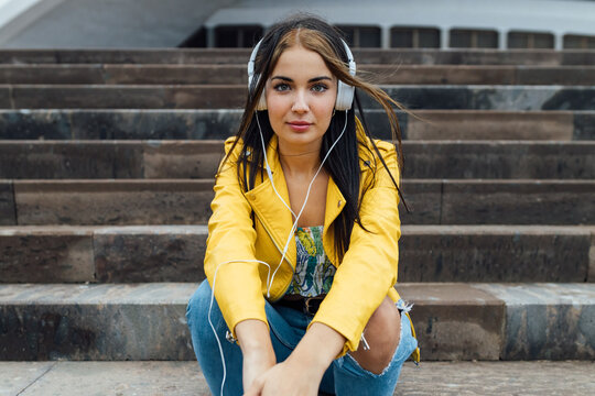 Low angle of young female in trendy jacket and ripped jeans looking at camera while sitting on stairs near urban building and listening to music through headphones