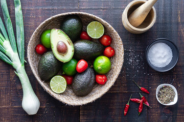 Top view bowl with fresh avocado and lime with tomatoes placed on wooden table with green onion and spices for traditional Mexican guacamole recipe
